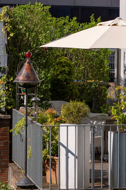 A small terrace garden featuring a variety of lush green plants and trees. In the foreground, there is a rectangular planter box with a large, leafy shrub, surrounded by pebbles inside the pot. The we