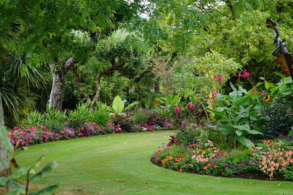 A lush, well-maintained garden features a curved, neatly mowed lawn bordered by vibrant flower beds filled with colorful blooms such as pink, orange, and purple flowers. Tall leafy shrubs and perennia