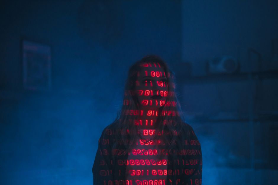 A person standing in a dark, indoor environment with their face and upper body illuminated by red binary code projection. The individual has long hair, and the digital numbers create a pattern across